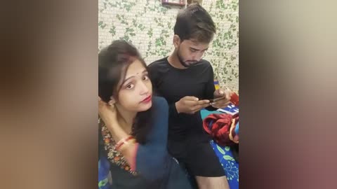 Media: Video of a young woman with dark hair and fair skin in a blue blouse, sitting on a bed with a patterned blanket, looking at a phone. Beside her, a young man in black shorts and shirt holds a phone, also looking at it.