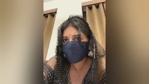 Media: Video of a woman with dark skin, wearing a black face mask, dark hair, and a sheer black polka-dot blouse, in a room with beige curtains.