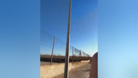 Media: Video of a barren, sunlit prison yard with a high chain-link fence, concrete walls, and a clear blue sky. The scene is desolate, emphasizing the stark, confined environment.