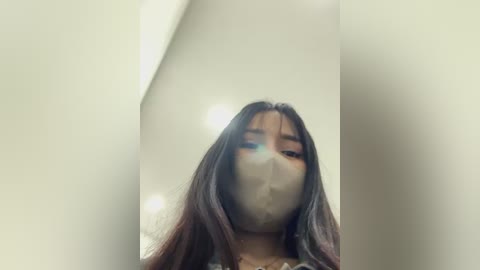 Media: Video of a young woman with long, dark hair and light skin, wearing a surgical mask, standing in a dimly lit room with white walls and a soft, blurred background.