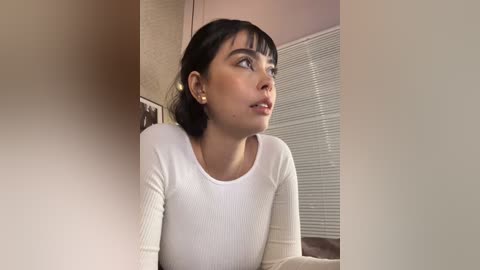 Media: Video of a young Asian woman with medium skin tone, short black hair, and bangs, wearing a white ribbed long-sleeve shirt. She gazes thoughtfully at a window with closed blinds. Background includes beige walls and a framed picture.