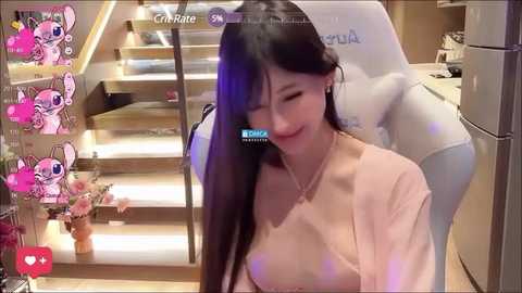 Media: Video of an Asian woman with long black hair, wearing a light pink blouse, sitting on a white chair. Background features a modern staircase and kitchen area.