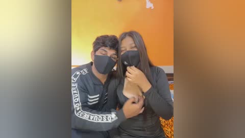 Media: Video of a South Asian couple with medium brown skin, wearing black face masks, standing in an orange room. The woman, with long black hair, has her breast exposed, while the man, with curly black hair, hugs her.