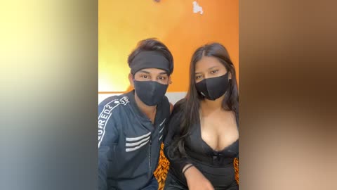 Media: Video of two South Asian women with medium skin tones, wearing black face masks and matching black jackets with white stripes. One has a low-cut black top, revealing cleavage. Background features an orange wall and a partial view of a colorful patterned blanket.