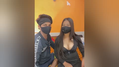 Media: Video of two South Asian women, one with medium-length black hair, the other with long black hair, both wearing black face masks and dark hoodies, sitting on a bed with orange walls and a colorful patterned blanket.