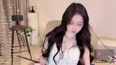 Media: Video of a young Asian woman with long black hair, wearing a white lace top, sitting at a wooden desk in a minimalistic room with beige curtains and a white chair.