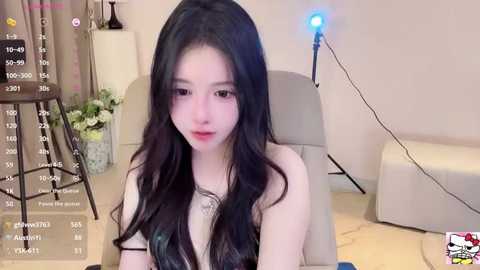 Media: Video of a young East Asian woman with long black hair, fair skin, and a slim build, sitting in a beige chair in a modern living room with a white lamp and potted plants.