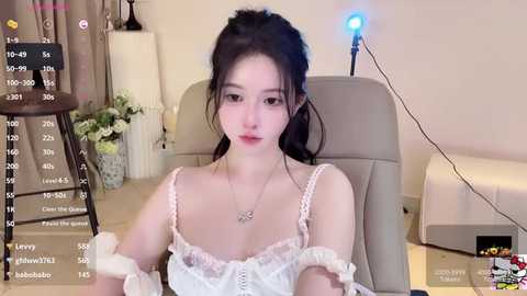 Media: A video of an Asian woman with fair skin, black hair in a ponytail, wearing a white off-shoulder dress, seated in a beige chair. The background features a potted plant, a lamp, and a \"Livestream\" overlay.