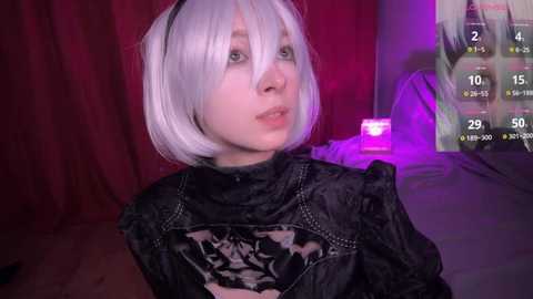 Media: Video of a pale-skinned woman with short, platinum blonde hair, wearing a black, high-necked dress with a floral pattern. She has a blank expression. Background includes a red curtain and a purple-lit screen displaying game stats.