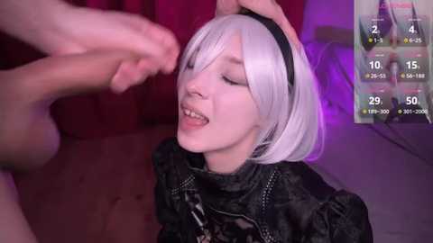 Media: A video of a young woman with a pale complexion, white bob haircut, and a black headband, smiling while a man's hand touches her head. She wears a black, textured jacket.