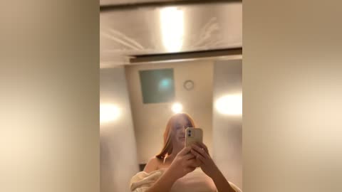 Media: A blurry selfie video of a light-skinned woman with long, straight hair, taken in a dimly lit, modern bathroom with beige walls and recessed lights. She is wearing a beige robe and holding a smartphone.