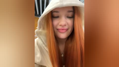 Media: Video of a young woman with long, vibrant orange hair, wearing a white hoodie, smiling. Background includes blurred, warm-toned walls.