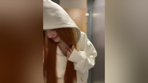 Media: Video of a woman with long, straight, reddish-brown hair wearing a white hoodie, hiding her face behind the hoodie's hood. She stands in a dimly lit, narrow hallway with beige walls.