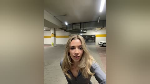 Media: Video of a blonde woman in a gray ribbed sweater, standing in a dimly lit, beige-walled parking garage with overhead lights and parked cars in the background.