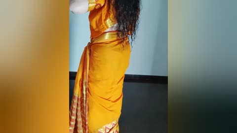 Media: Video of a woman in a vibrant yellow saree with red and gold patterns, standing with her back to the camera. Her long, curly dark hair cascades down her back.