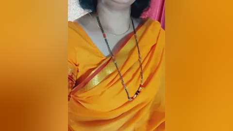 Media: A video of a person in a vibrant yellow sari with a gold border, adorned with a long, beaded necklace. The background is a solid orange color, and the person's face is not visible.