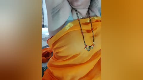 Media: Video of a woman's torso in an orange sari, showcasing intricate golden border and colorful jewelry. She wears a blue top and a red chain necklace, with a blurred background featuring a bed and a black object.