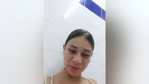 Media: A video of a young woman with dark hair pulled back, wearing a beige top, in a white-tiled bathroom with blue accents. She has a contemplative expression.