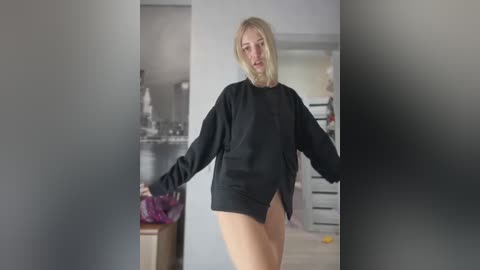 Media: Video of a blonde woman in a black, oversized sweater, standing in a modern, minimalist room with gray walls and minimal furniture.