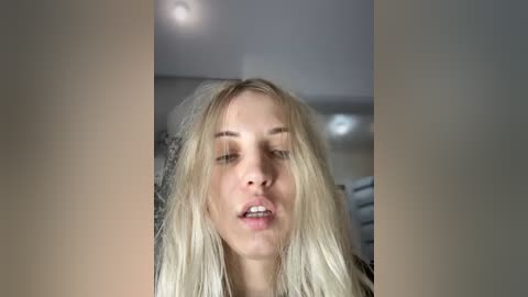 Media: Video of a young, fair-skinned woman with long, straight blonde hair, slightly parted lips, and closed eyes. She appears to be in a room with recessed lighting and blurred background furniture.