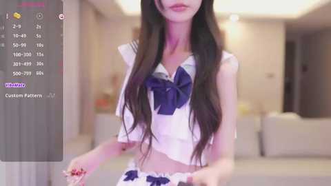 Media: A video of a slender Asian woman with long, dark hair, wearing a white sailor-style crop top with a blue bow, standing in a modern, dimly-lit living room.