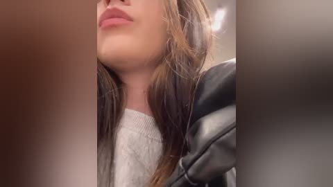 Media: Video of a young woman with light skin and long brown hair, wearing a white sweater and a black leather jacket, seen from a side angle, blurred background.