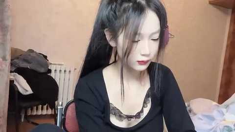 Media: Video of a young Asian woman with long black hair, wearing a black top and lacy bra, sitting in a cluttered bedroom with a bed and clothes.