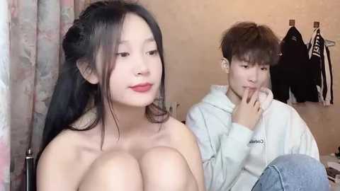 Media: Video of a topless East Asian woman with long black hair and red lipstick, standing next to a young man in a hoodie. Background features a beige wall with hanging jackets.