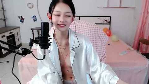 Media: Video of an East Asian woman with dark hair, wearing a white satin robe, smiling while singing into a microphone in a pink-checkered bedroom.