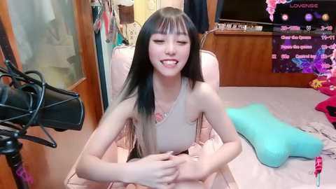 Media: Video of an East Asian woman with long black hair and fair skin, wearing a beige sleeveless top, smiling, sitting on a bed with turquoise pillow, surrounded by colorful bedding and clothes in a cluttered room.