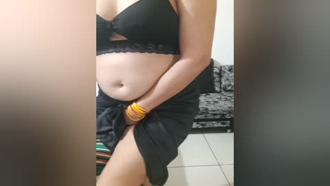 Media: Video of a light-skinned woman with a belly, wearing a black bra and a black skirt with a high slit, holding her skirt, wearing gold bangles. Background shows a black couch and colorful rug.