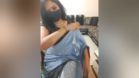 Media: A video captures a woman in a living room, wearing a blue shirt, black mask, and gray pants, adjusting her shirt. The background features a black and white patterned couch and a white coffee table.