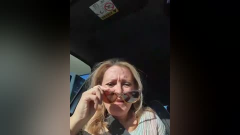 Media: A video of a middle-aged woman with blonde hair, wearing sunglasses, and a light-colored blouse, seated in a car with a \"No Smoking\" sign above her head.