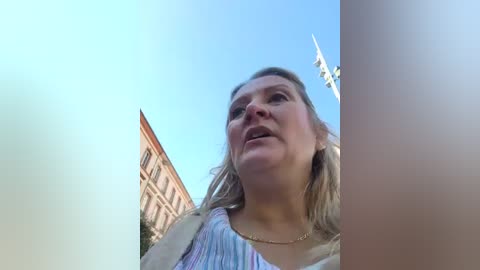 Media: A video featuring a middle-aged woman with blonde hair, wearing a colorful top, captured from a low angle, partially obscured by a tall building and a clear blue sky.