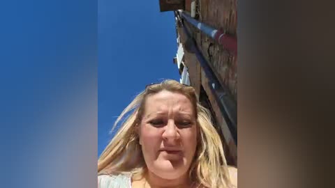 Media: Video of a middle-aged woman with blonde hair, wearing a light-colored top, standing against a clear blue sky and a brick building with a metal gutter, looking slightly distressed.