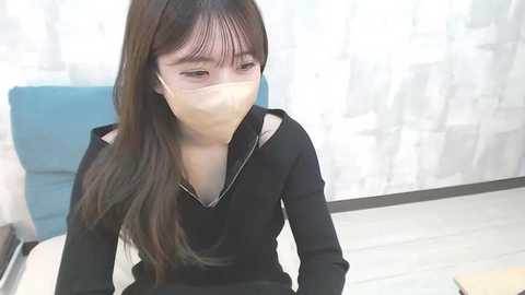 Media: Video of an Asian woman with long, straight black hair, wearing a black jacket, a face mask, and sitting in a room with light-colored curtains and a blue sofa.