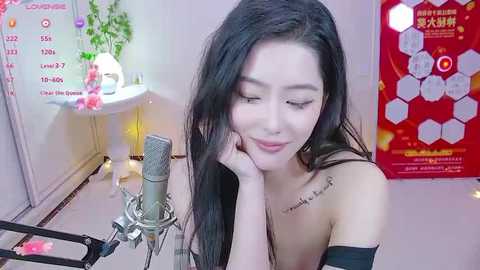 Media: Video of an Asian woman with long black hair, wearing a beige off-shoulder top, smiling while resting her chin on her hand, microphone close-up. Background includes a whiteboard, red poster, and green plants.