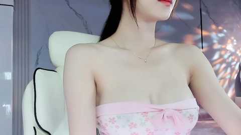 Media: Video of an East Asian woman with pale skin, wearing a strapless, pink floral dress with a bow, sitting on a white chair. Background features a modern, gray and white interior with a large, abstract, colorful painting.