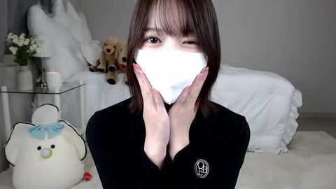 Media: Video of an Asian woman with shoulder-length brown hair, wearing a black long-sleeve top, white face mask, and a plush toy. She stands in a softly lit bedroom with white bedding, a stuffed dog, and a table with a vase of flowers.