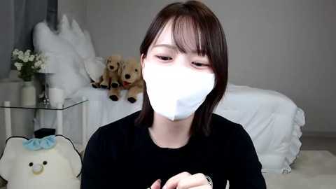 Media: Video of a young woman with straight, shoulder-length brown hair, wearing a white surgical mask, black top, in a minimalist bedroom with white bedding, stuffed animals, and a glass table.