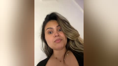 Media: Video of a young Latina woman with medium brown skin, long wavy hair, and a confident smile. She's wearing a black top, standing in front of a plain white background.