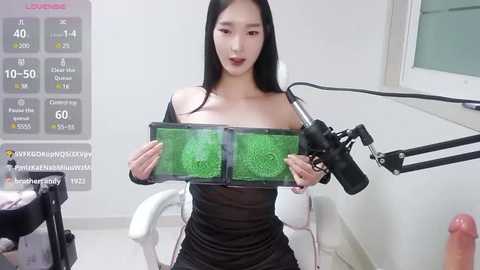 Media: A video of an East Asian woman with long black hair, fair skin, and medium-sized breasts, wearing a tight black dress, holding a green-screen prop. The setting is a studio with a white wall and microphone.