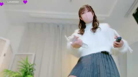 Media: Video of a young woman with long brown hair, wearing a white turtleneck and gray pleated skirt, holding a smartphone, indoors with a potted plant and white curtains in the background.