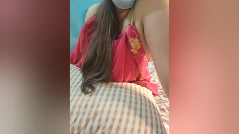 Media: Video of a woman with long brown hair wearing a red floral blouse and gray face mask, lying on a patterned bedspread, blurred background.