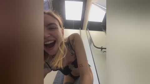 Media: A video captures a blonde woman with fair skin, wearing a black bra, laughing ecstatically, with a tattoo on her left arm. She is in a brightly lit, modern bathroom with white walls and a glass shower enclosure.