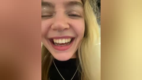 Media: A close-up video of a young woman with light skin, long blonde hair, and a happy expression, wearing a black turtleneck and a silver necklace.