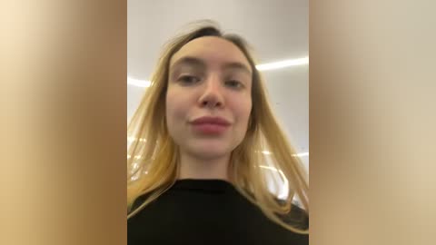 Media: Video of a young Caucasian woman with long blonde hair, wearing a black top, standing in a modern room with a white ceiling and soft lighting.