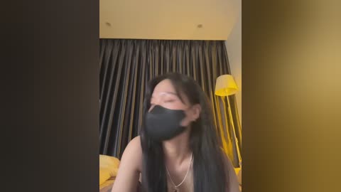 Media: Video of a young East Asian woman with long black hair, wearing a black face mask and black top, standing in a dimly lit room with dark curtains and a yellow lamp.