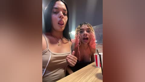 Media: Video of two young women, one with long black hair, the other with curly pink hair, playfully sticking out their tongues at the camera. They are dressed in casual tops, seated at a wooden table in a dimly lit restaurant.