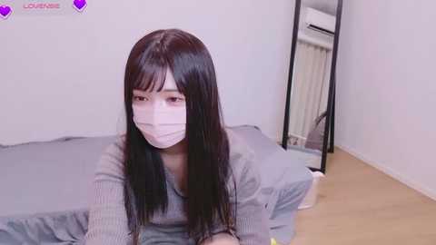 Media: Video of a young East Asian woman with long, straight black hair, wearing a face mask, grey sweater, and leaning on a grey bed in a simple, sparsely furnished room with a mirror, white wall, and wooden floor.